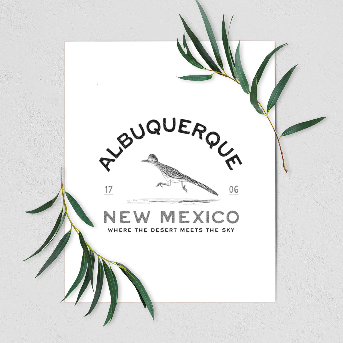 Art print of a black ink roadrunner with the words Albuquerque New Mexico, 1706, Where the Desert Meets the Sky, shown with eucalyptus leaves.