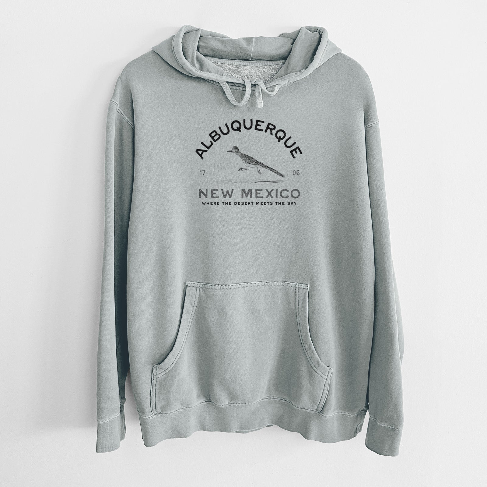 Sage green pigment dyed hoodie laid flat, printed with a black ink roadrunner with the words Albuquerque New Mexico, 1706, Where the Desert Meets the Sky.