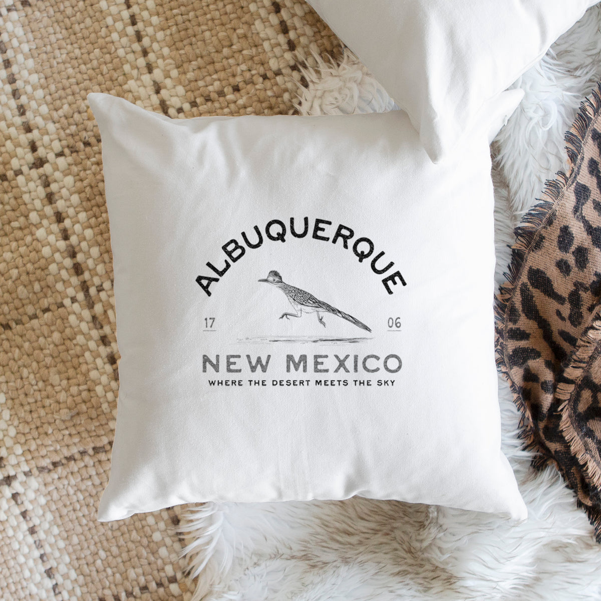 White pillow cover printed with a black ink roadrunner with the words Albuquerque New Mexico, 1706, Where the Desert Meets the Sky, on a woven chair.