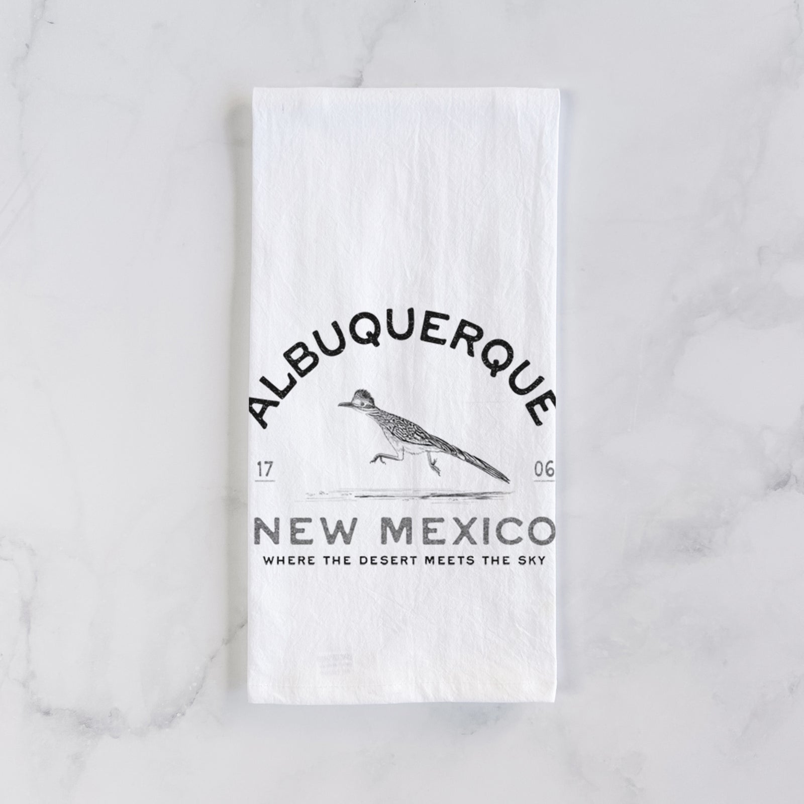 White cotton tea towel printed with a black ink roadrunner with the words Albuquerque New Mexico, 1706, Where the Desert Meets the Sky, on a marble counter beside a round wooden cutting board and greenery.