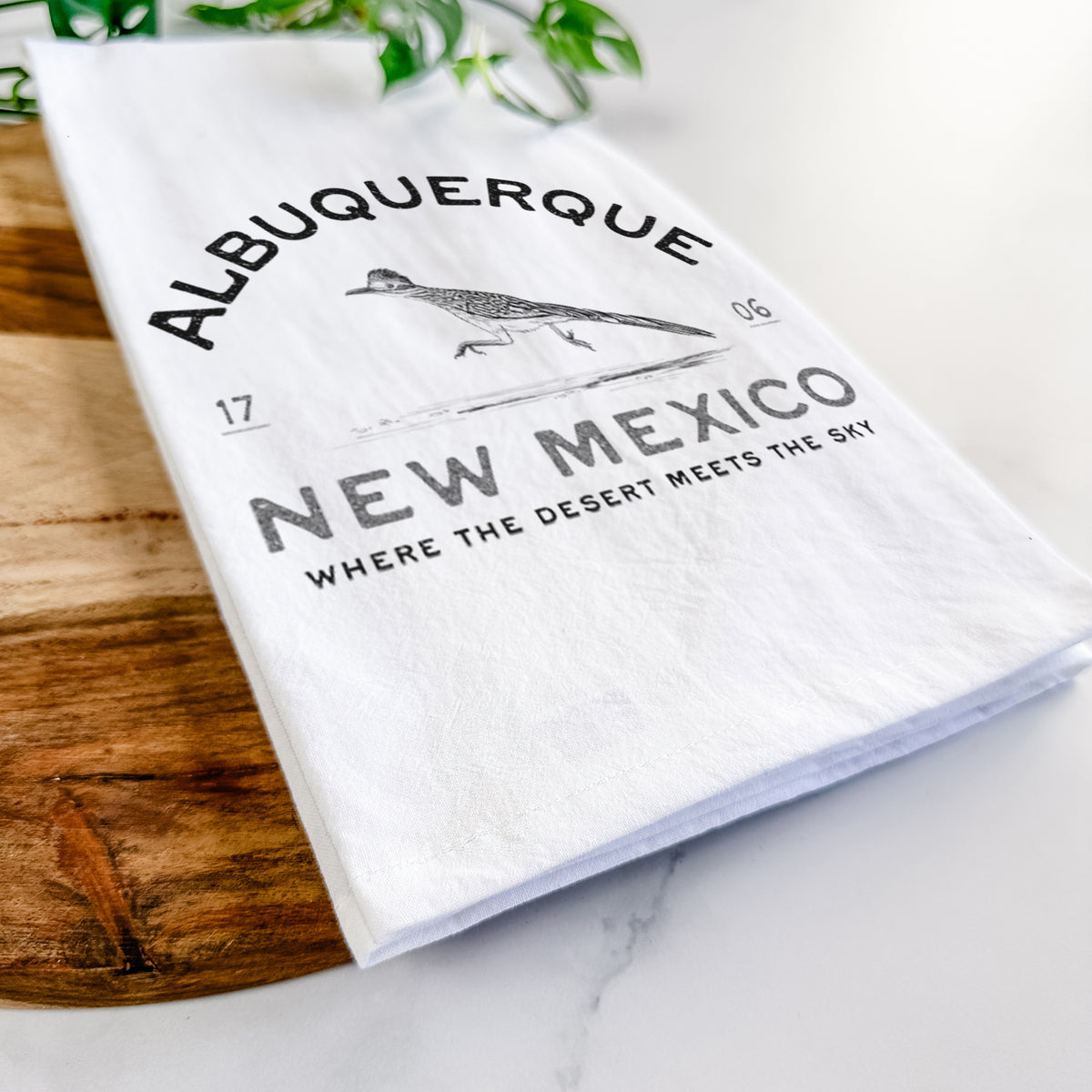 Close-up of a folded white tea towel on a wooden board, showing a black ink roadrunner with the words Albuquerque New Mexico, 1706, Where the Desert Meets the Sky.