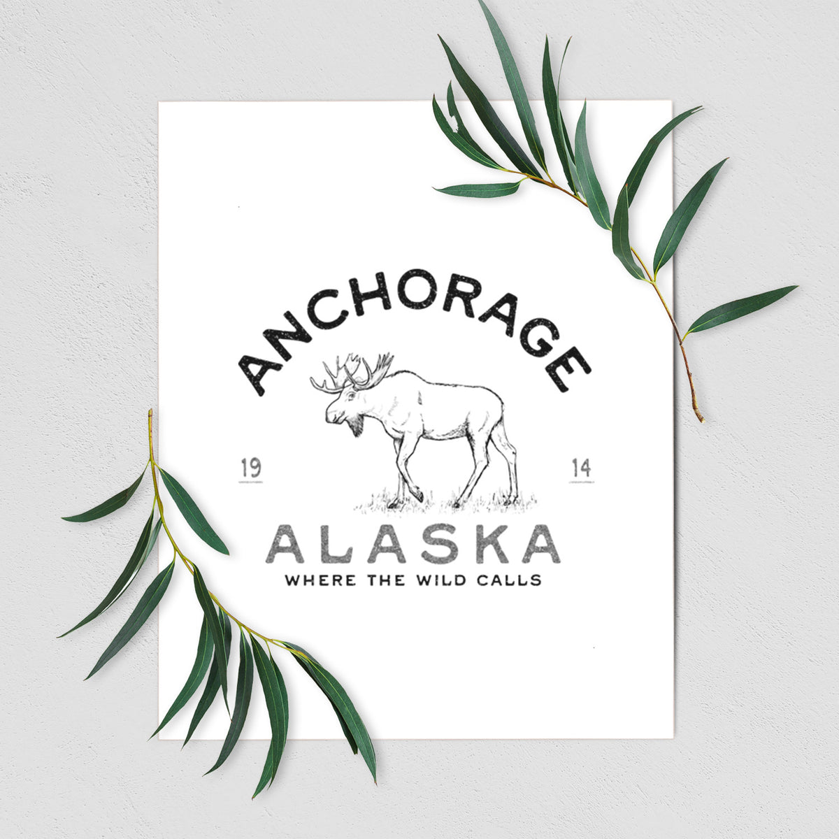 Art print of a black ink moose with the words Anchorage Alaska, 1914, Where the Wild Calls, shown with eucalyptus leaves.