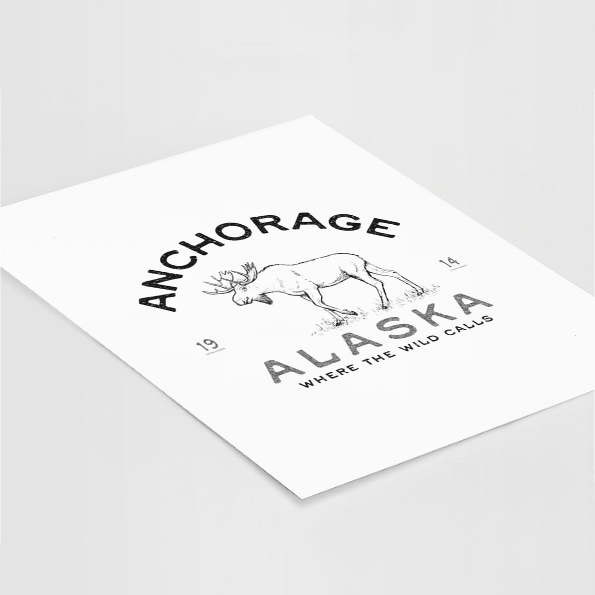 Art print of a black ink moose with the words Anchorage Alaska, 1914, Where the Wild Calls, lying at an angle on a white surface.