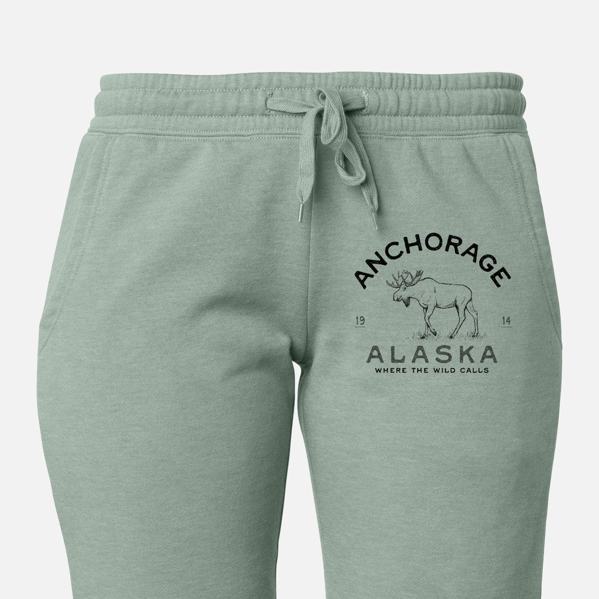Front of sage green Cali Wave sweatpants, printed with a black ink moose with the words Anchorage Alaska, 1914, Where the Wild Calls on the leg.