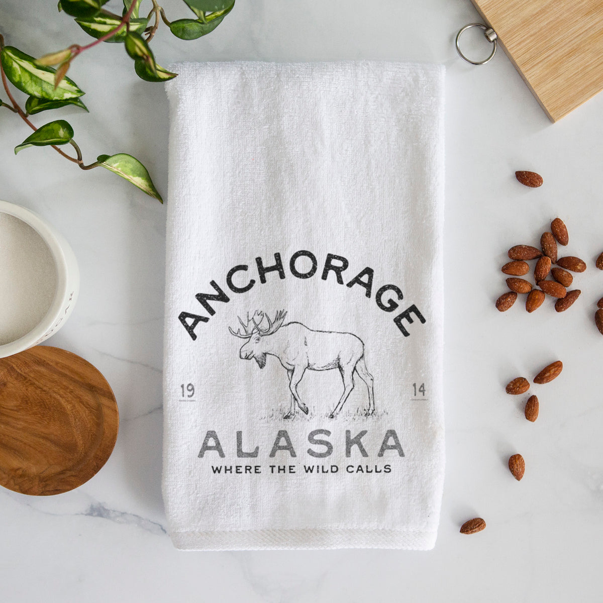 White hand towel printed with a black ink moose with the words Anchorage Alaska, 1914, Where the Wild Calls.