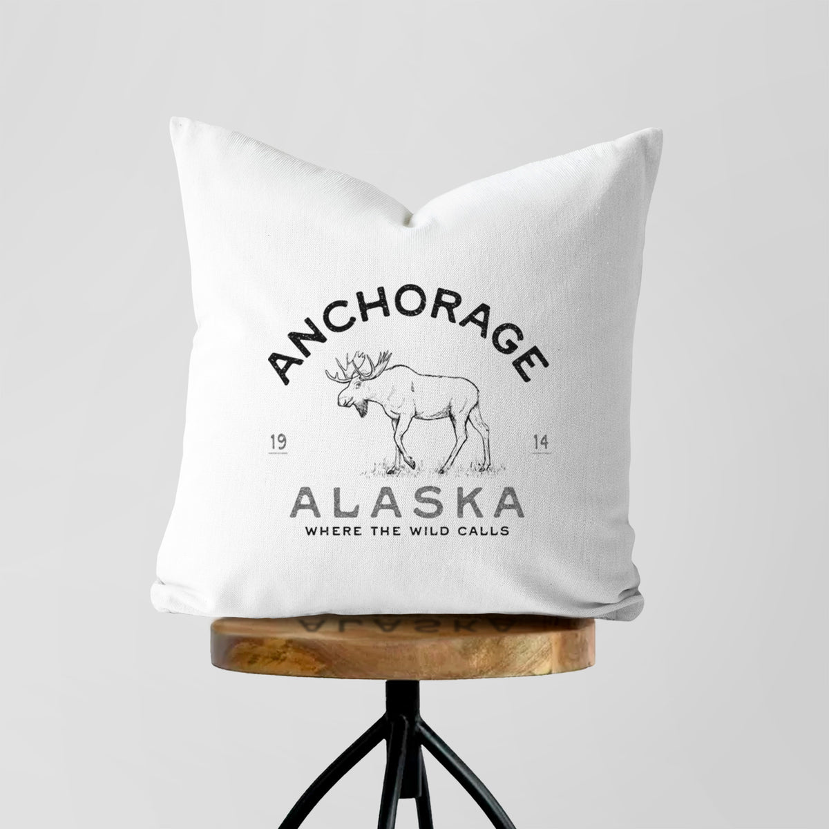 White pillow cover printed with a black ink moose with the words Anchorage Alaska, 1914, Where the Wild Calls, on a wooden stool.