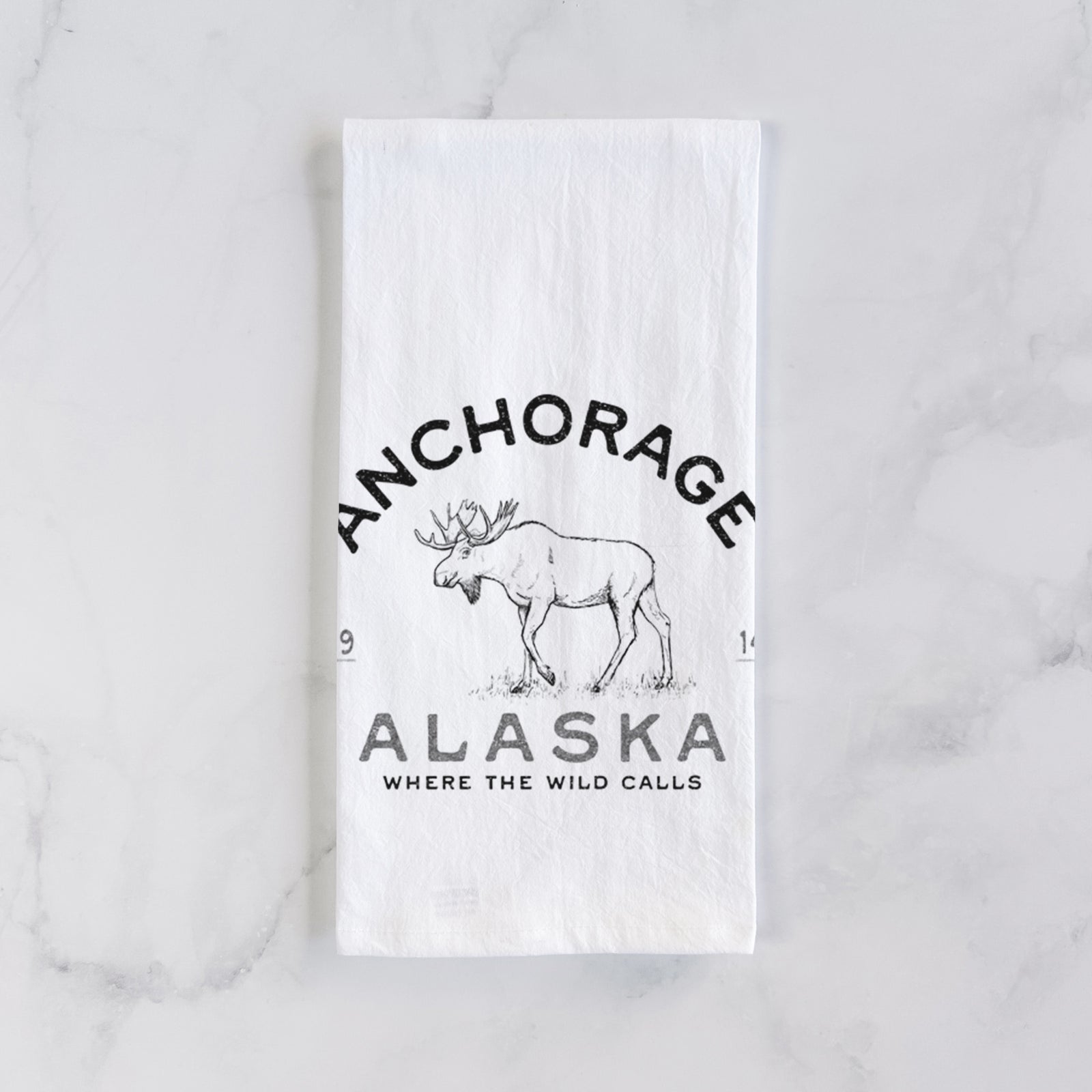 White cotton tea towel printed with a black ink moose with the words Anchorage Alaska, 1914, Where the Wild Calls, on a marble counter beside a round wooden cutting board and greenery.