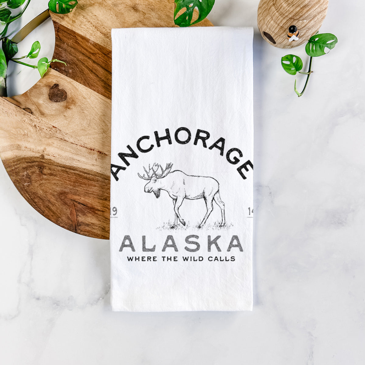 White cotton tea towel printed with a black ink moose with the words Anchorage Alaska, 1914, Where the Wild Calls, on a marble counter beside a round wooden cutting board and greenery.
