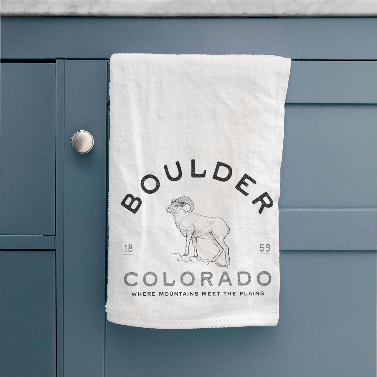 White hand towel printed with a black ink bighorn sheep with the words Boulder Colorado, Where Mountains Meet the Plains, 1859, hanging on a cabinet door.