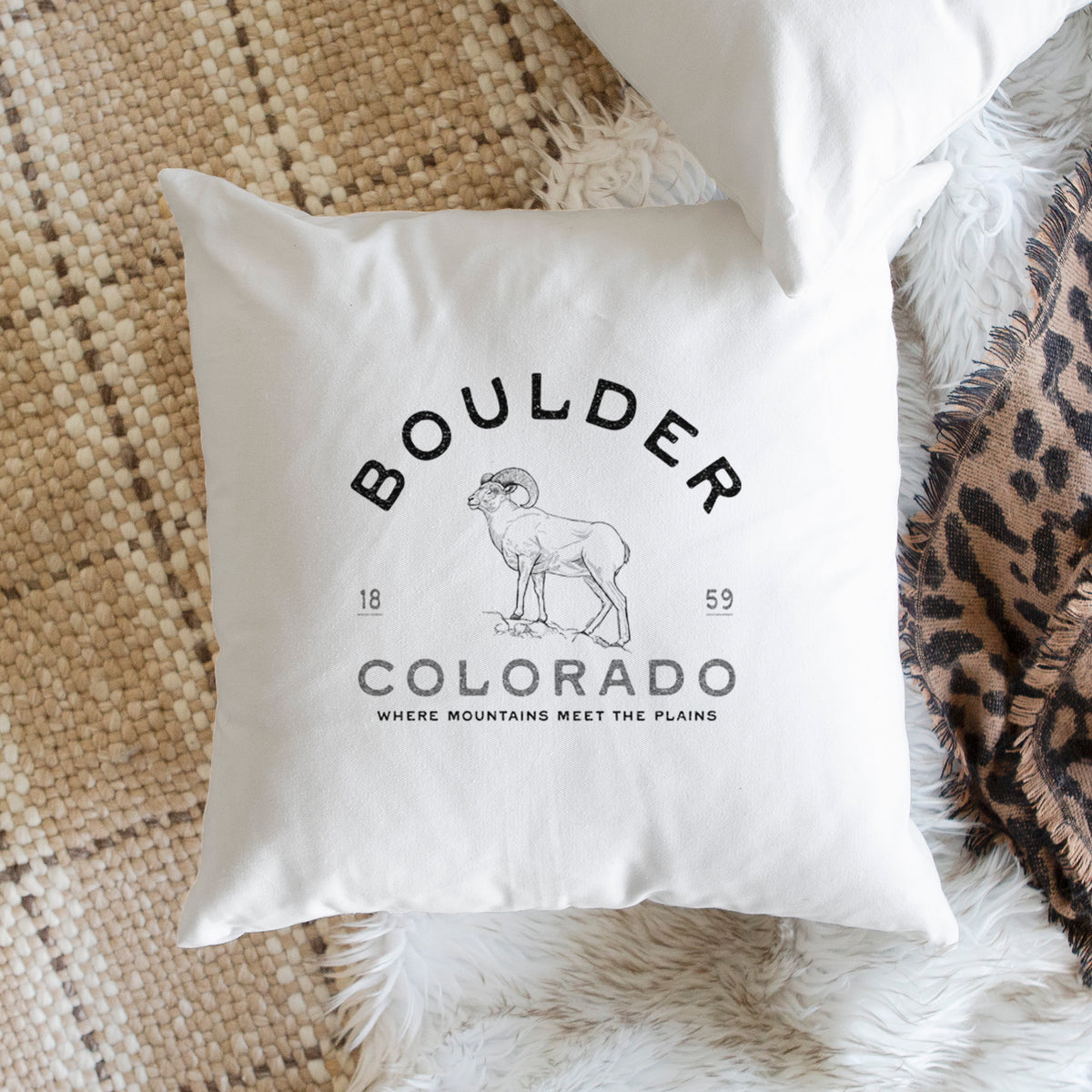 White pillow cover printed with a black ink bighorn sheep with the words Boulder Colorado, Where Mountains Meet the Plains, 1859, on a woven chair.