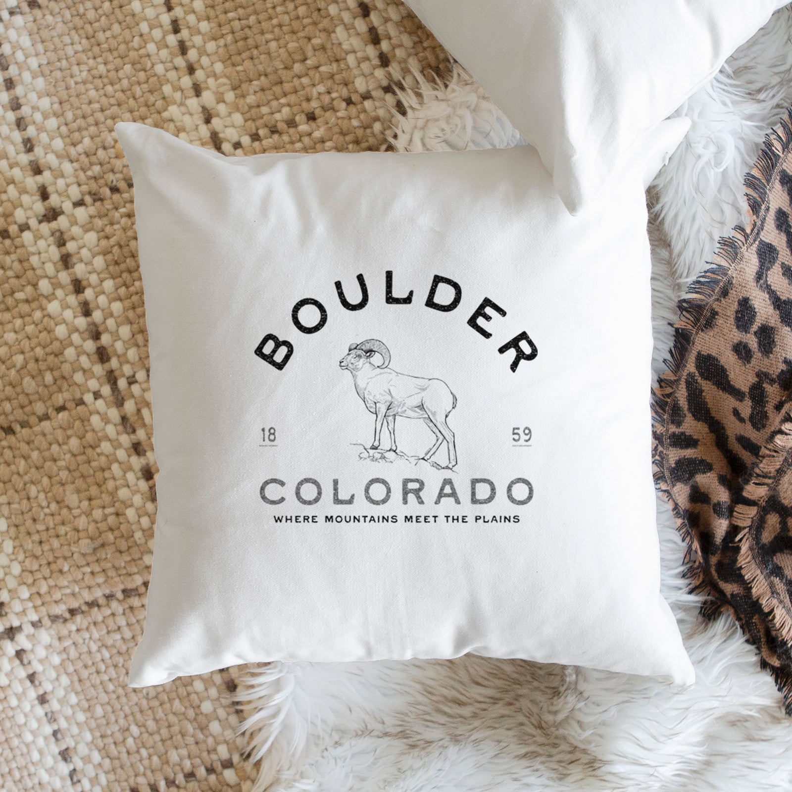 White pillow cover printed with a black ink bighorn sheep with the words Boulder Colorado, Where Mountains Meet the Plains, 1859, on a bench beside plants.