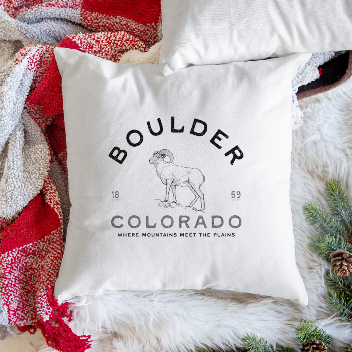 White pillow cover printed with a black ink bighorn sheep with the words Boulder Colorado, Where Mountains Meet the Plains, 1859, on a woven chair.