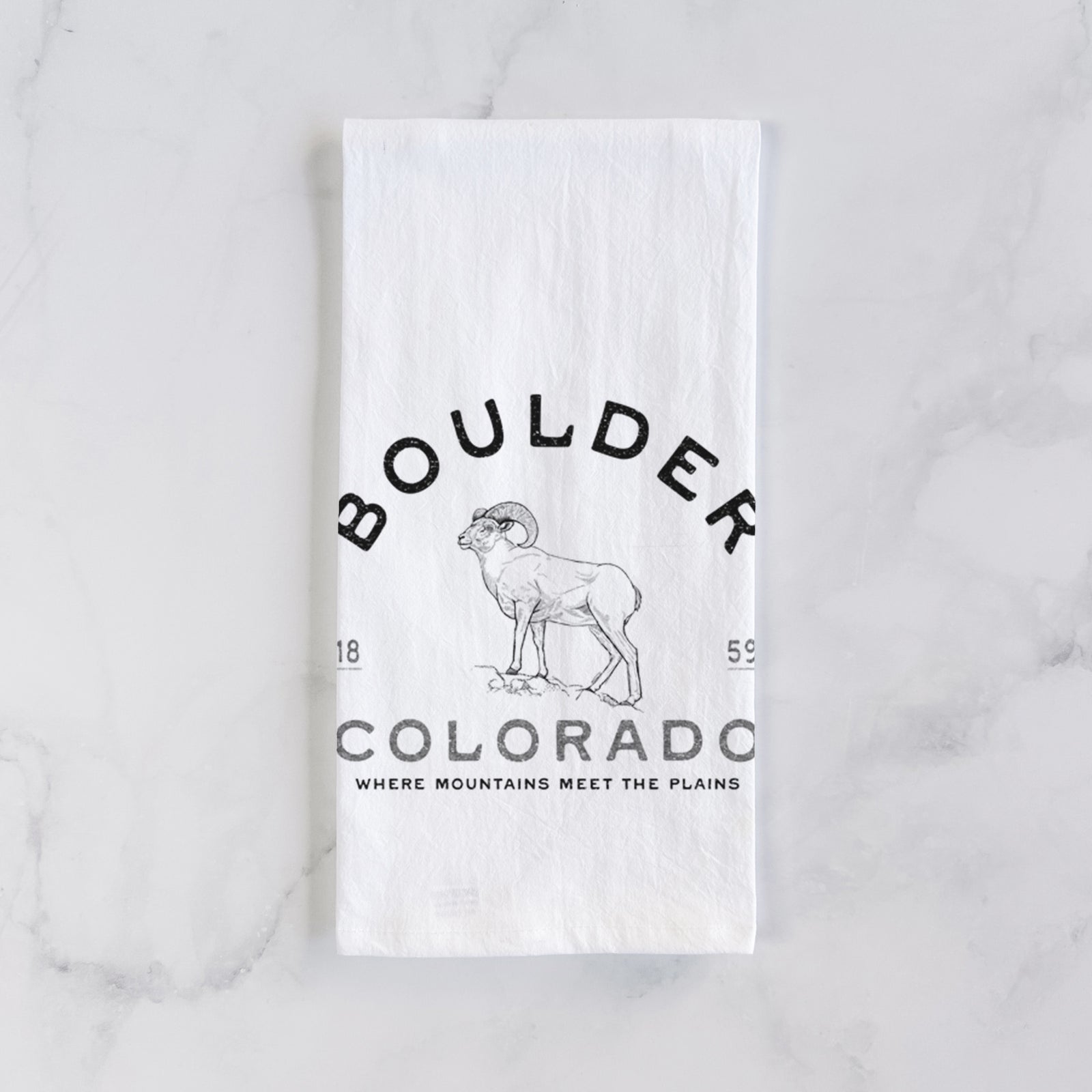 White cotton tea towel printed with a black ink bighorn sheep with the words Boulder Colorado, Where Mountains Meet the Plains, 1859, on a marble counter beside a round wooden cutting board and greenery.