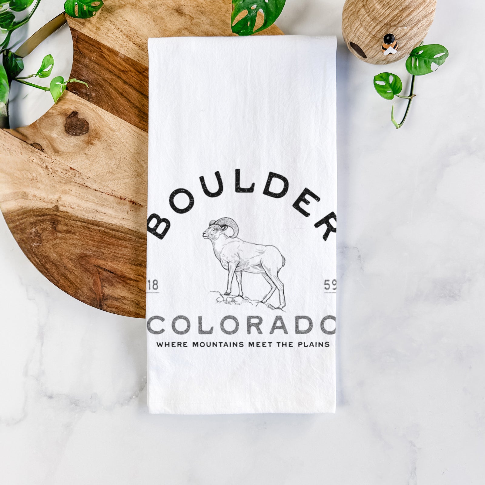 White cotton tea towel printed with a black ink bighorn sheep with the words Boulder Colorado, Where Mountains Meet the Plains, 1859, on a marble counter beside a round wooden cutting board and greenery.