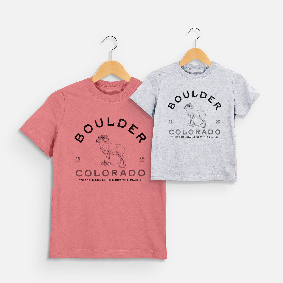 Two youth t-shirts in different colors on hangers, printed with a black ink bighorn sheep with the words Boulder Colorado, Where Mountains Meet the Plains, 1859.