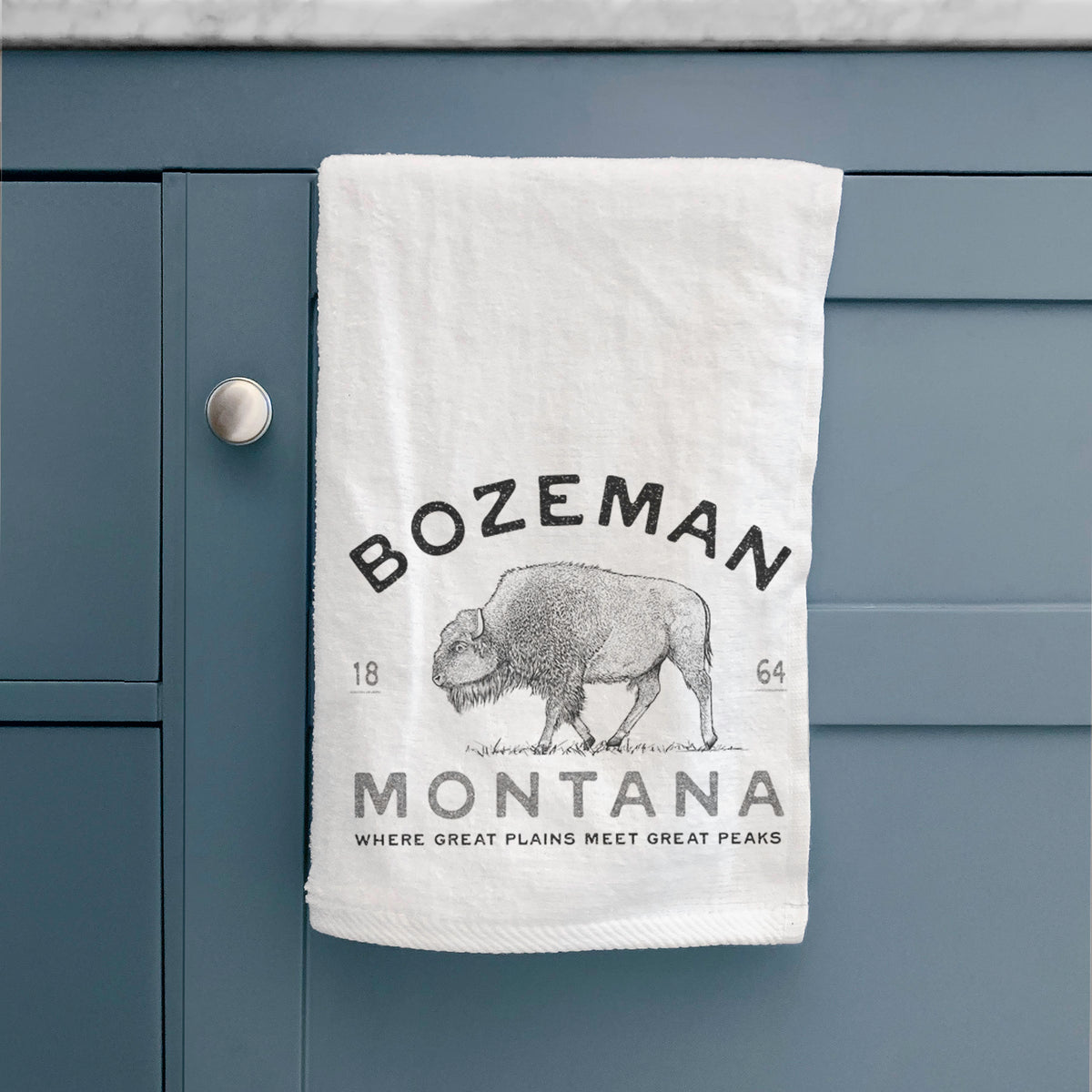 White hand towel printed with a black ink bison with the words Bozeman Montana, 1864, Where Great Plains Meet Great Peaks, hanging on a cabinet door.