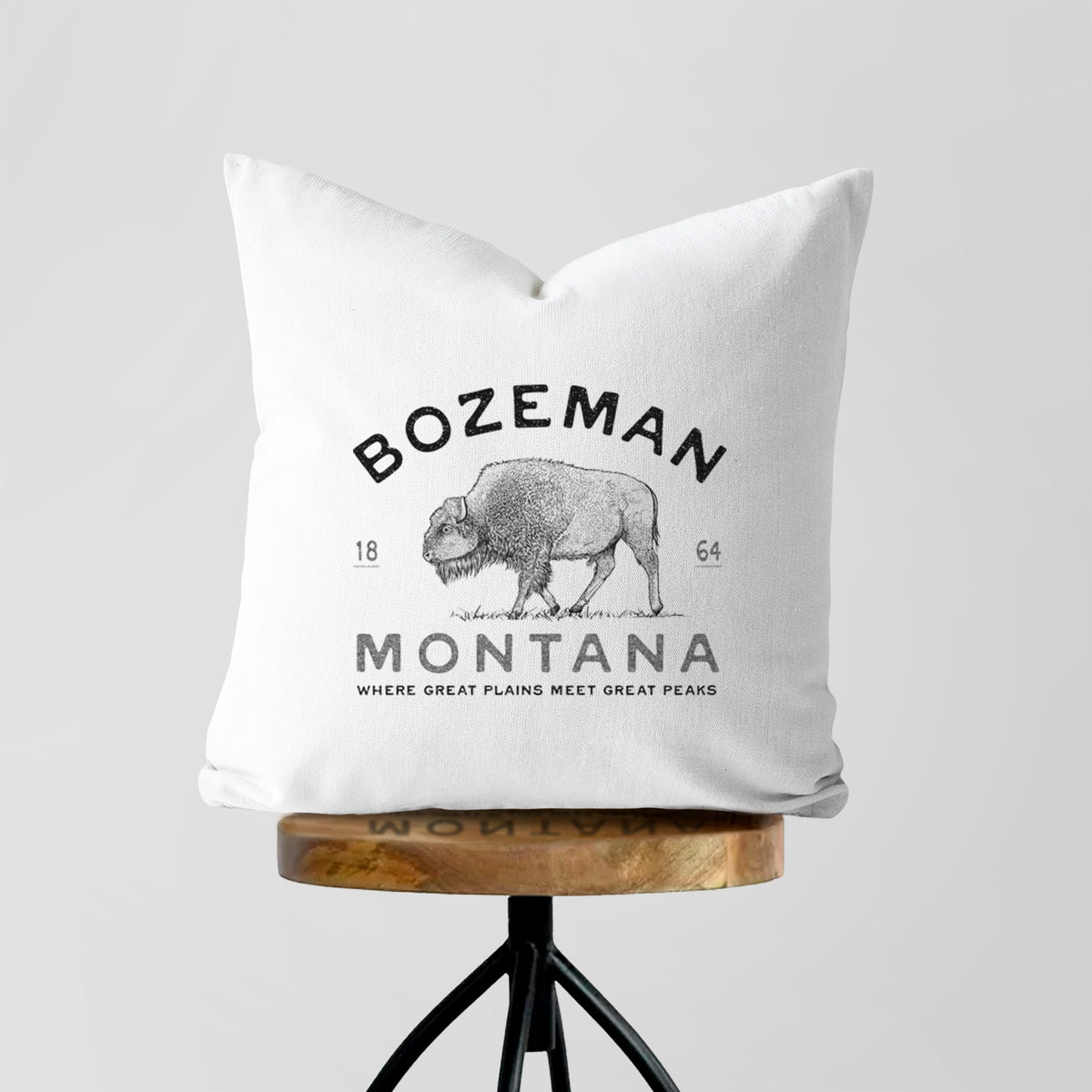 White pillow cover printed with a black ink bison with the words Bozeman Montana, 1864, Where Great Plains Meet Great Peaks, on a wooden stool.
