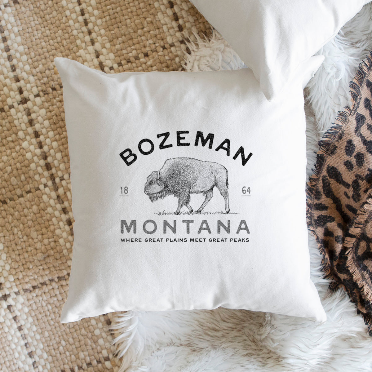 White pillow cover printed with a black ink bison with the words Bozeman Montana, 1864, Where Great Plains Meet Great Peaks, on a woven chair.