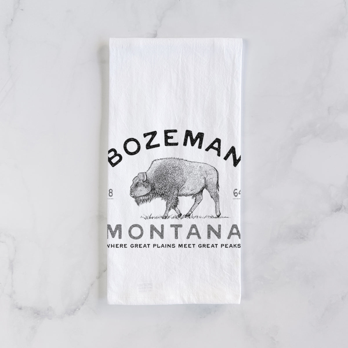 White cotton tea towel laid flat on marble, printed with a black ink bison with the words Bozeman Montana, 1864, Where Great Plains Meet Great Peaks.