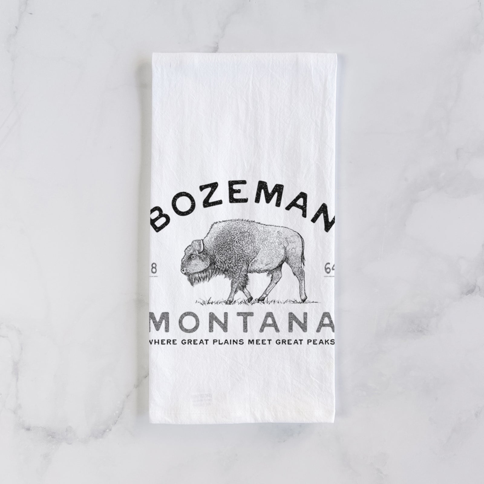 White cotton tea towel printed with a black ink bison with the words Bozeman Montana, 1864, Where Great Plains Meet Great Peaks, on a marble counter beside a round wooden cutting board and greenery.