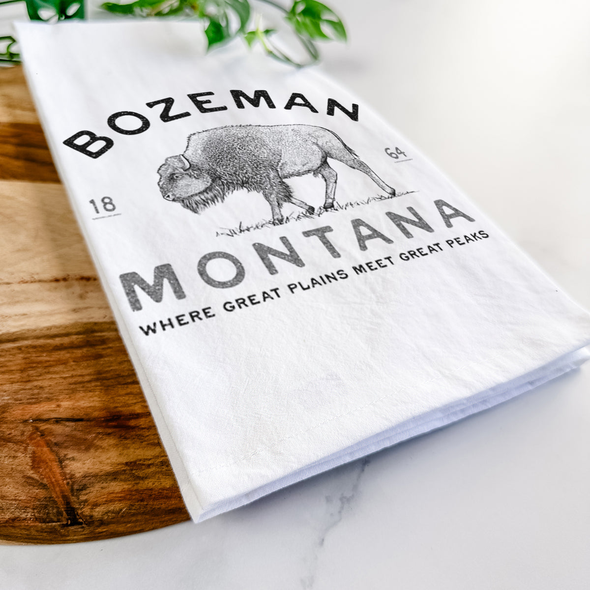 Close-up of a folded white tea towel on a wooden board, showing a black ink bison with the words Bozeman Montana, 1864, Where Great Plains Meet Great Peaks.