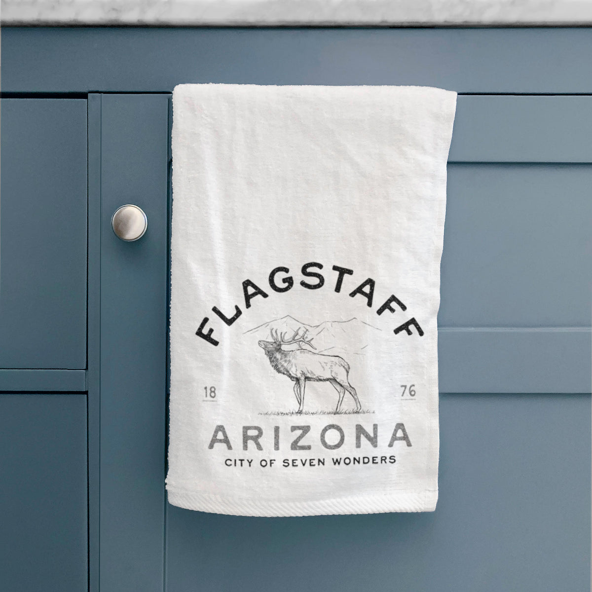 White hand towel printed with a black ink elk with the words Flagstaff Arizona, City of Seven Wonders, hanging on a cabinet door.