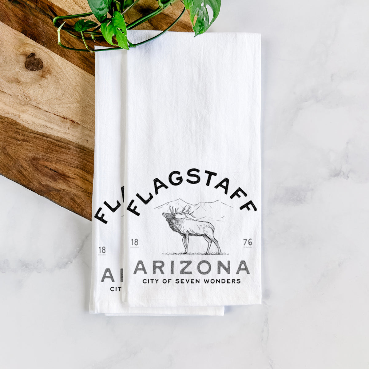 White cotton tea towel printed with a black ink elk with the words Flagstaff Arizona, City of Seven Wonders, on a marble counter with a wooden cutting board and greenery.