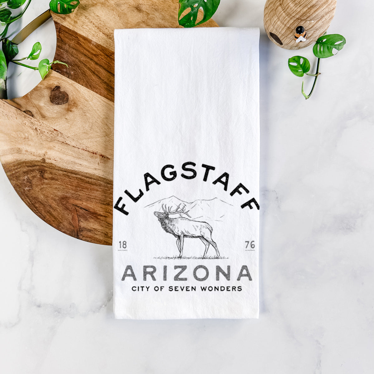 White cotton tea towel printed with a black ink elk with the words Flagstaff Arizona, City of Seven Wonders, on a marble counter beside a round wooden cutting board and greenery.