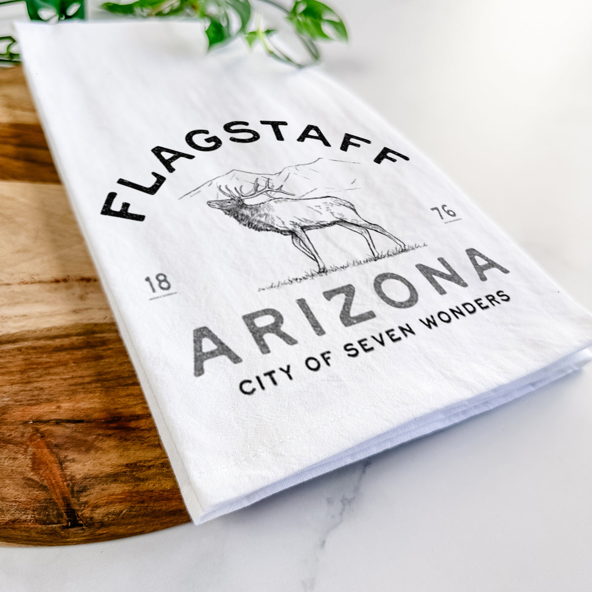 Close-up of a folded white tea towel on a wooden board, showing a black ink elk with the words Flagstaff Arizona, City of Seven Wonders.