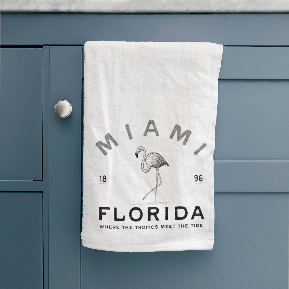 White hand towel printed with a black ink flamingo with the words Miami Florida, 1896, Where the Tropics Meet the Tide, hanging on a cabinet door.