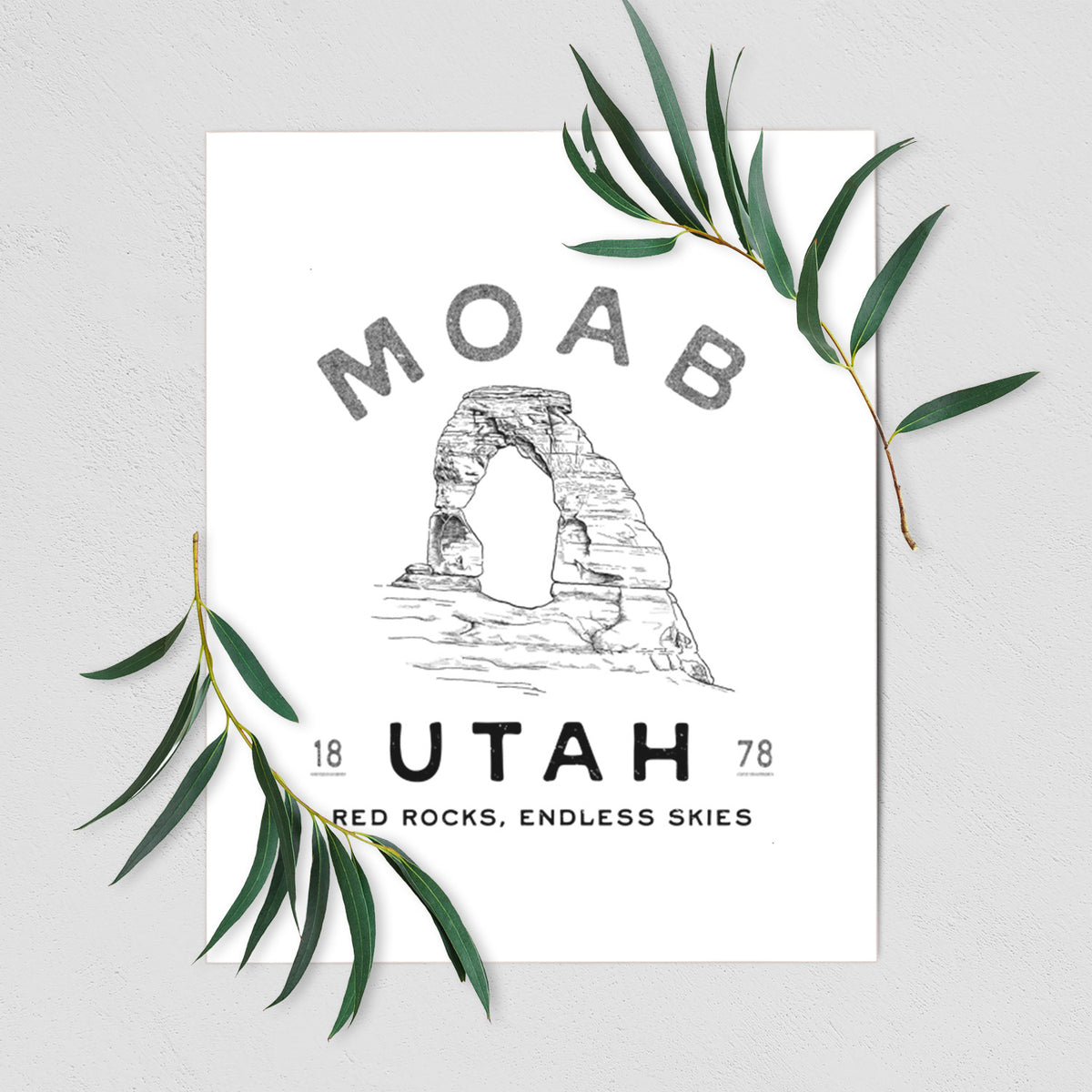 Art print of a black ink illustration of Delicate Arch with the words Moab Utah, 1878, shown with eucalyptus leaves.