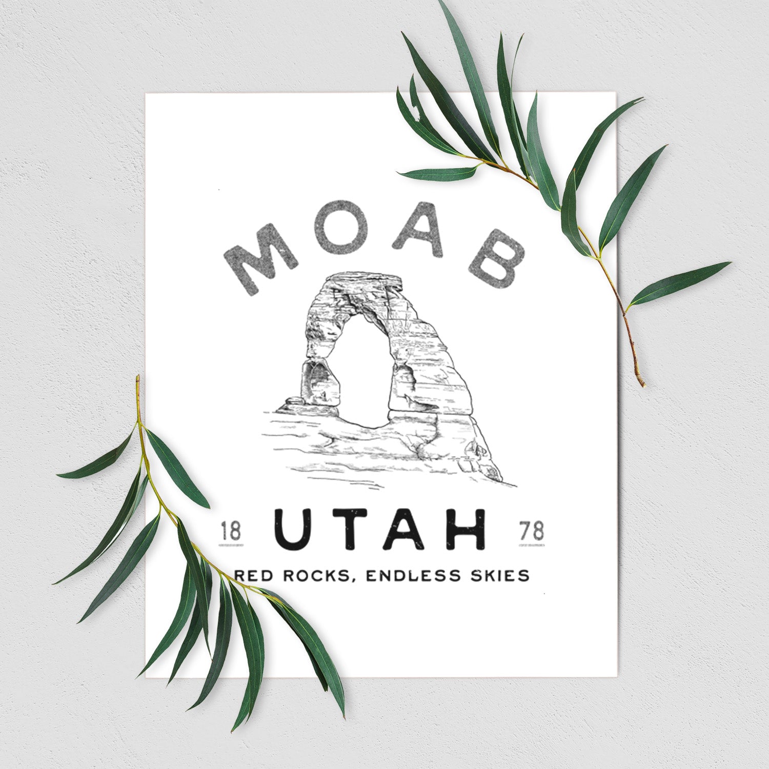 Art print of a black ink illustration of Delicate Arch with the words Moab Utah, 1878, shown with eucalyptus leaves.
