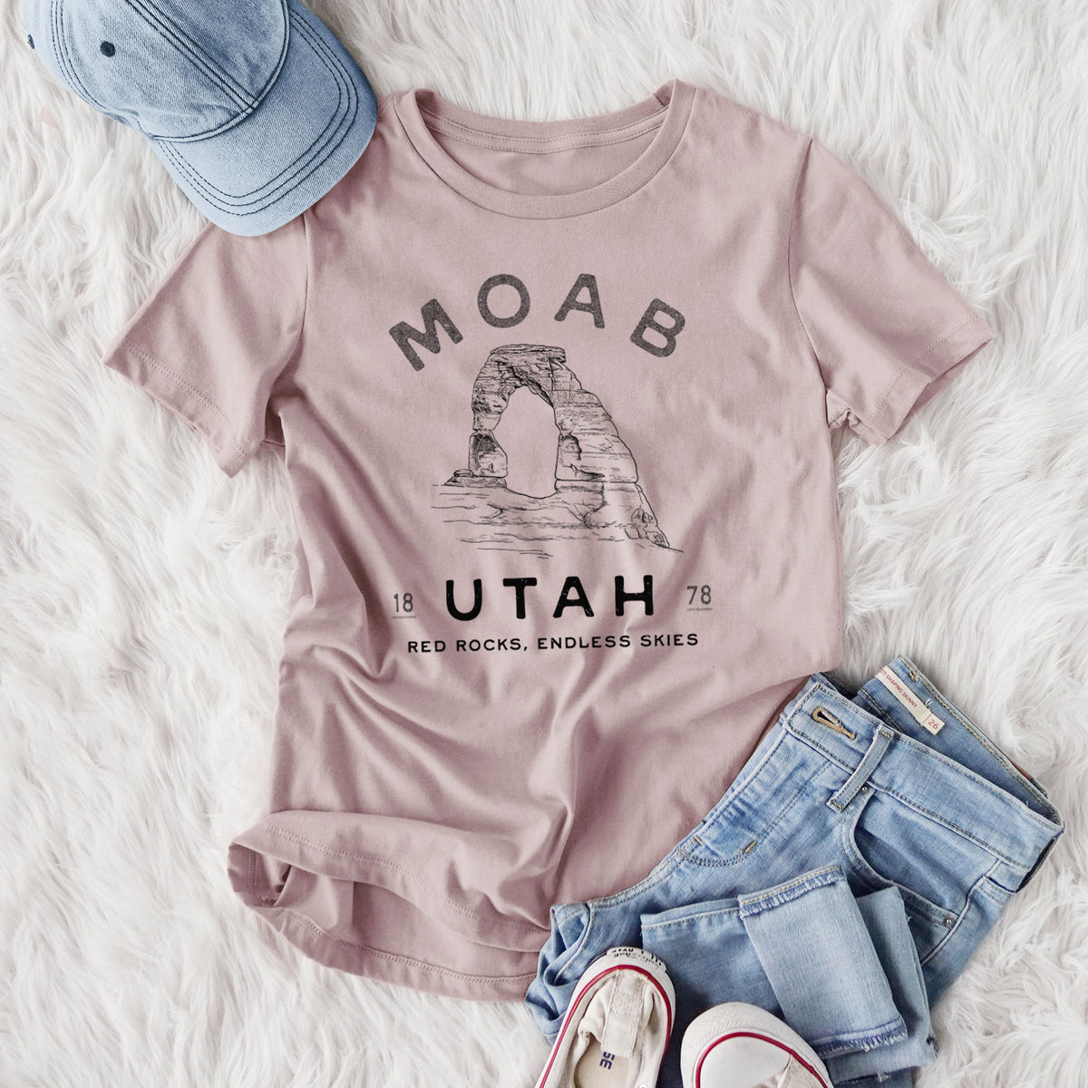 Women's relaxed fit cotton t-shirt printed with a black ink illustration of Delicate Arch with the words Moab Utah, 1878, styled flat on a white faux fur rug with jeans.