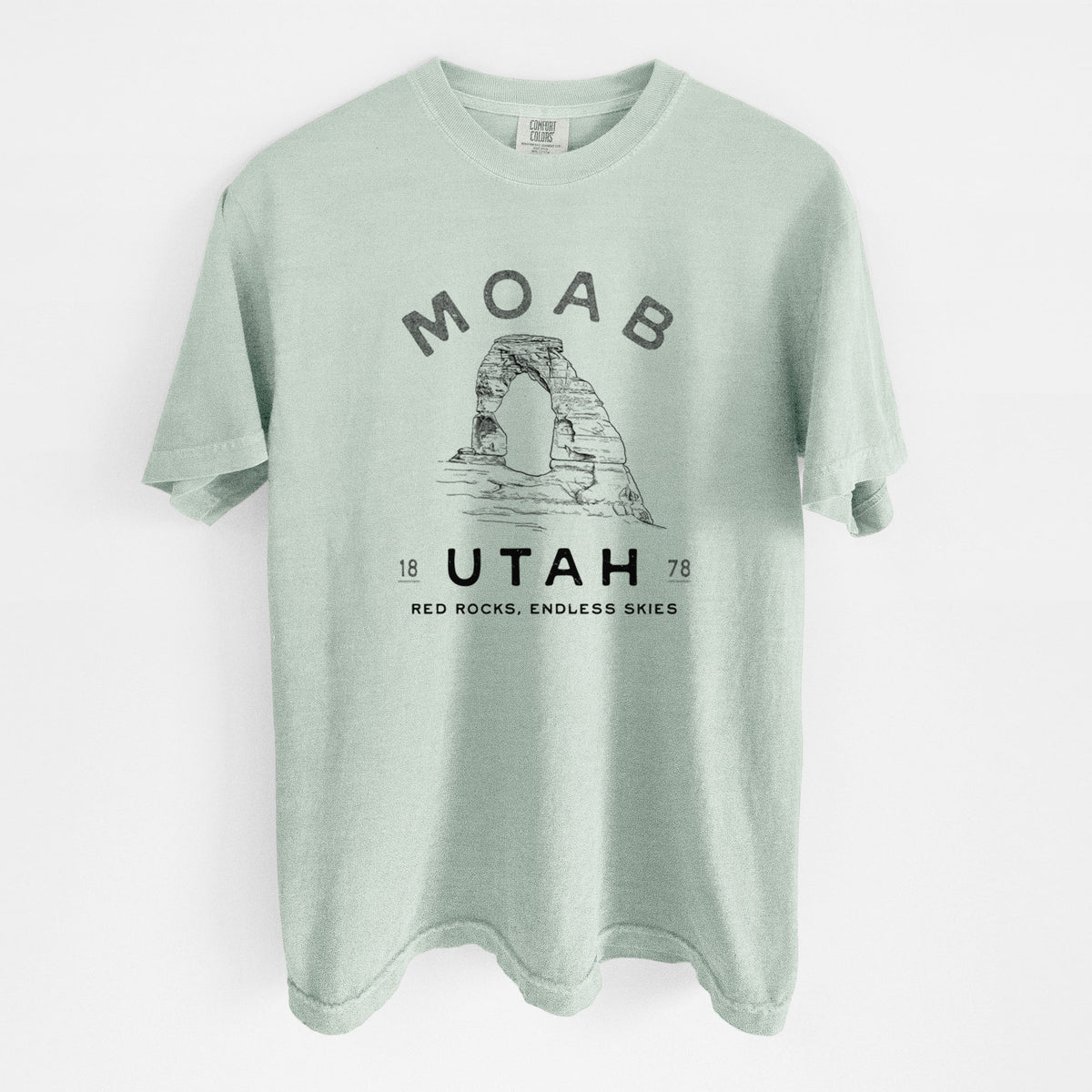 Bay green Comfort Colors t-shirt laid flat, printed with a black ink illustration of Delicate Arch with the words Moab Utah, 1878.