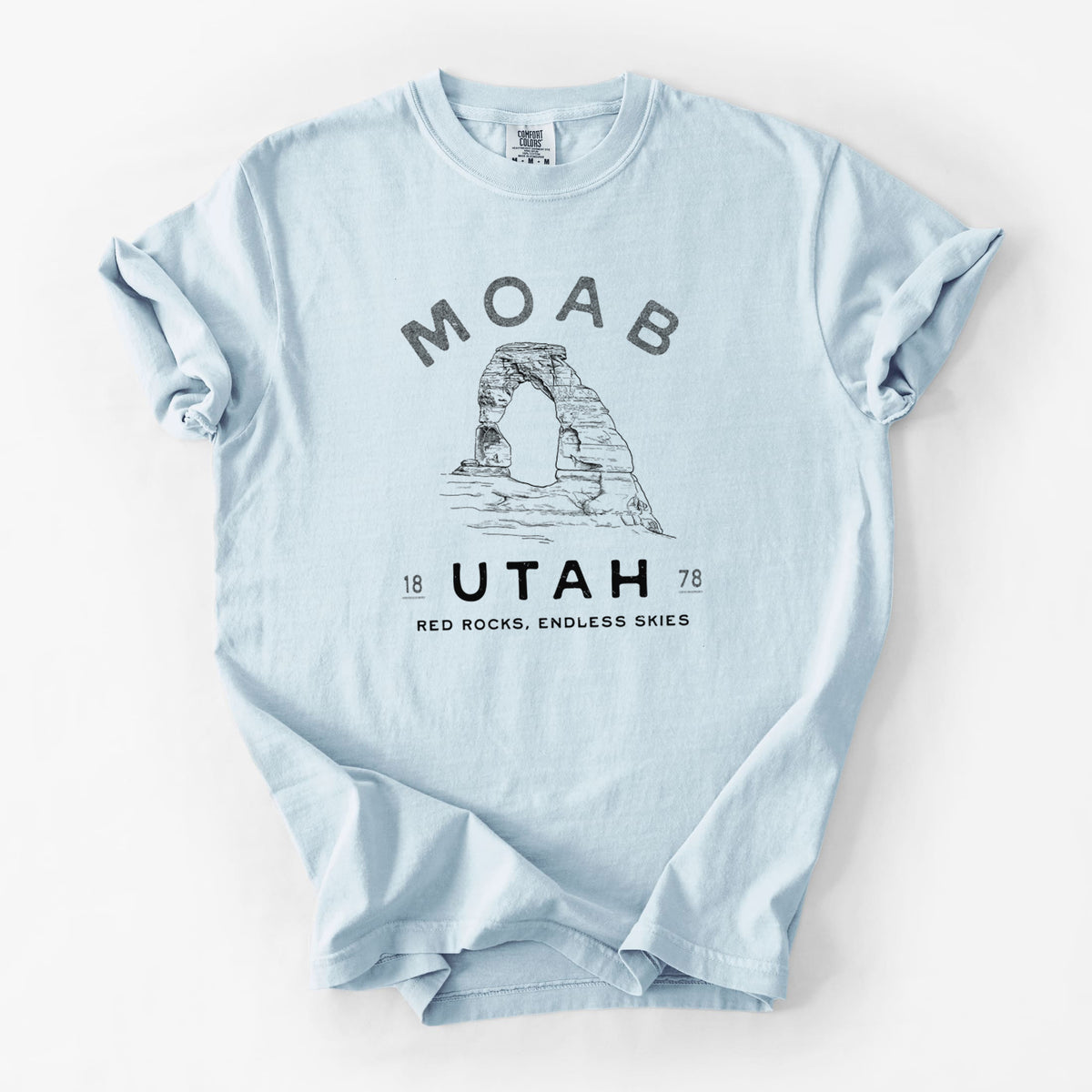 Chambray blue Comfort Colors t-shirt laid flat, printed with a black ink illustration of Delicate Arch with the words Moab Utah, 1878.