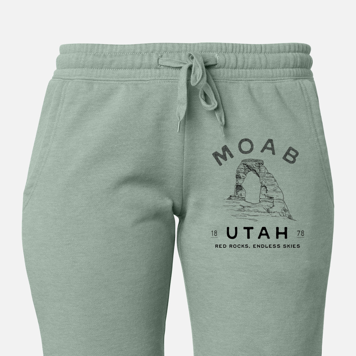 Front of sage green Cali Wave sweatpants, printed with a black ink illustration of Delicate Arch with the words Moab Utah, 1878 on the leg.