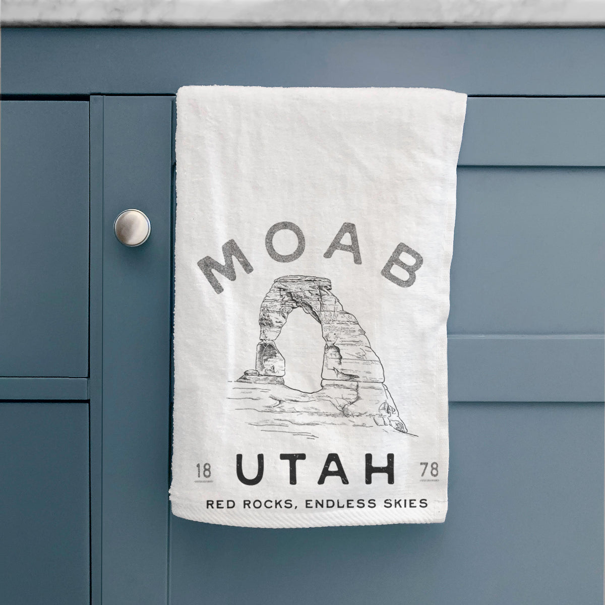 White hand towel printed with a black ink illustration of Delicate Arch with the words Moab Utah, 1878, hanging on a cabinet door.