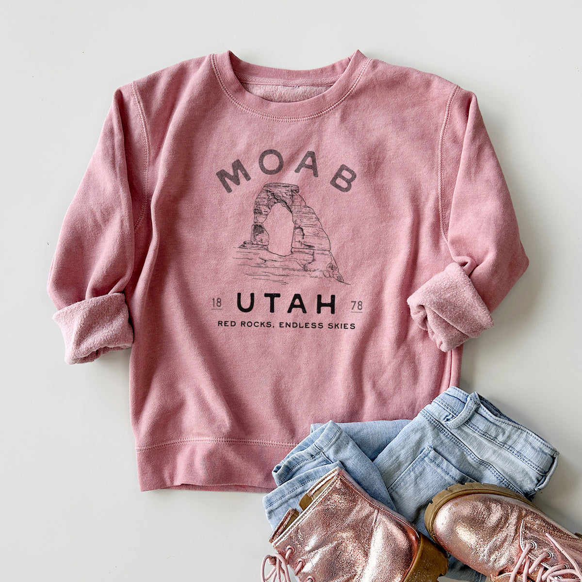 Youth crewneck sweatshirt printed with a black ink illustration of Delicate Arch with the words Moab Utah, 1878, styled flat with jeans and boots.