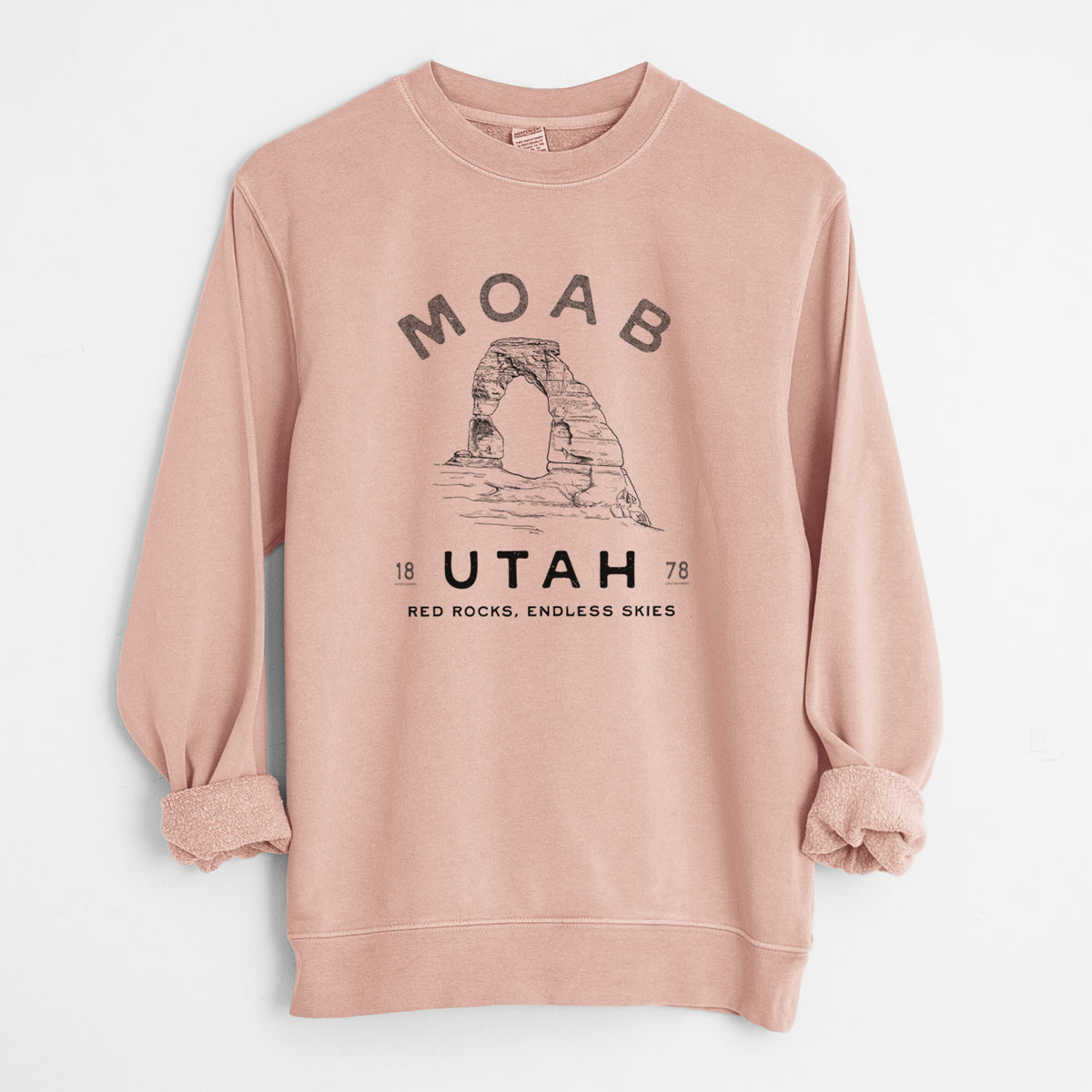 Dusty pink pigment dyed crewneck sweatshirt laid flat with the sleeves rolled, printed with a black ink illustration of Delicate Arch with the words Moab Utah, 1878.