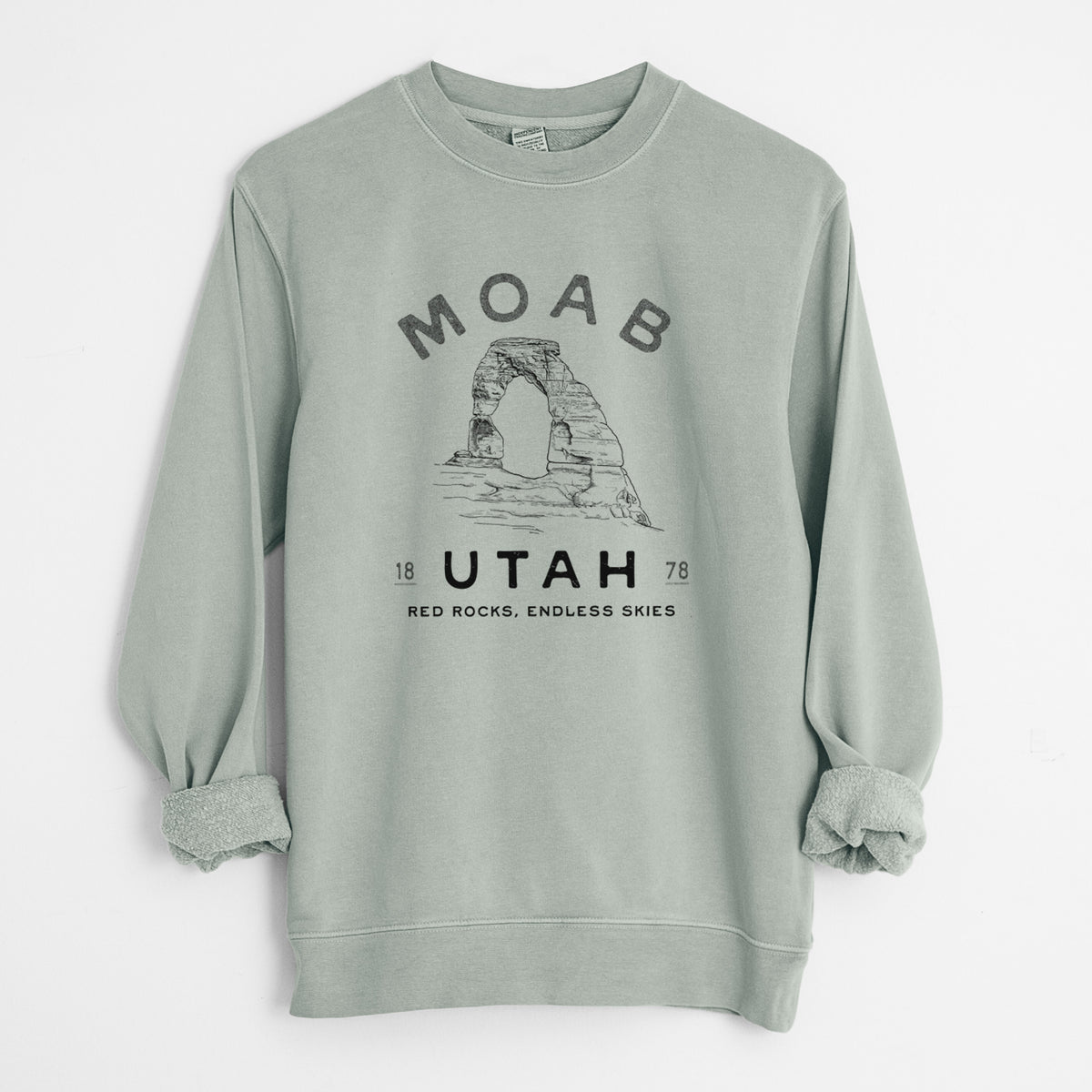 Sage green pigment dyed crewneck sweatshirt laid flat with the sleeves rolled, printed with a black ink illustration of Delicate Arch with the words Moab Utah, 1878.