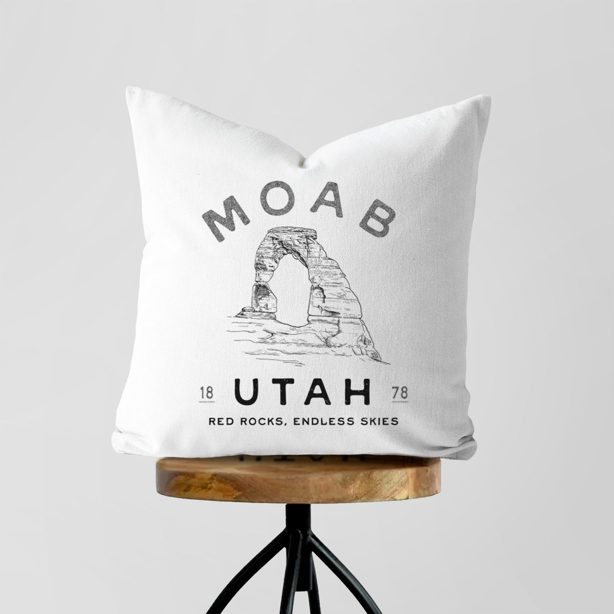 White pillow cover printed with a black ink illustration of Delicate Arch with the words Moab Utah, 1878, on a wooden stool.
