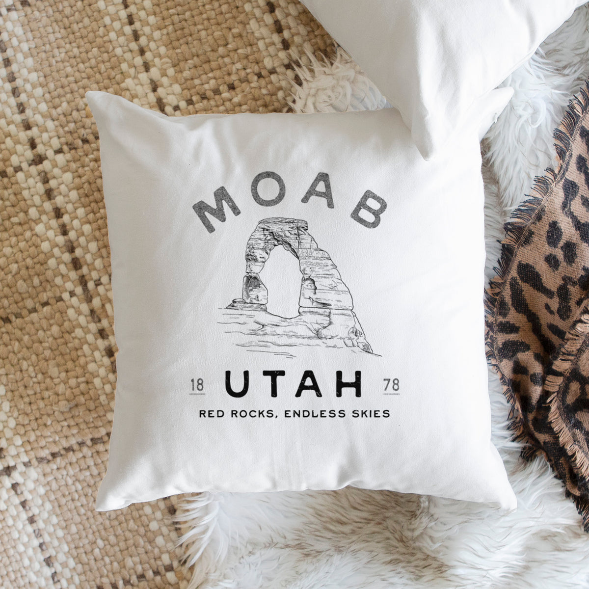 White pillow cover printed with a black ink illustration of Delicate Arch with the words Moab Utah, 1878, on a woven chair.