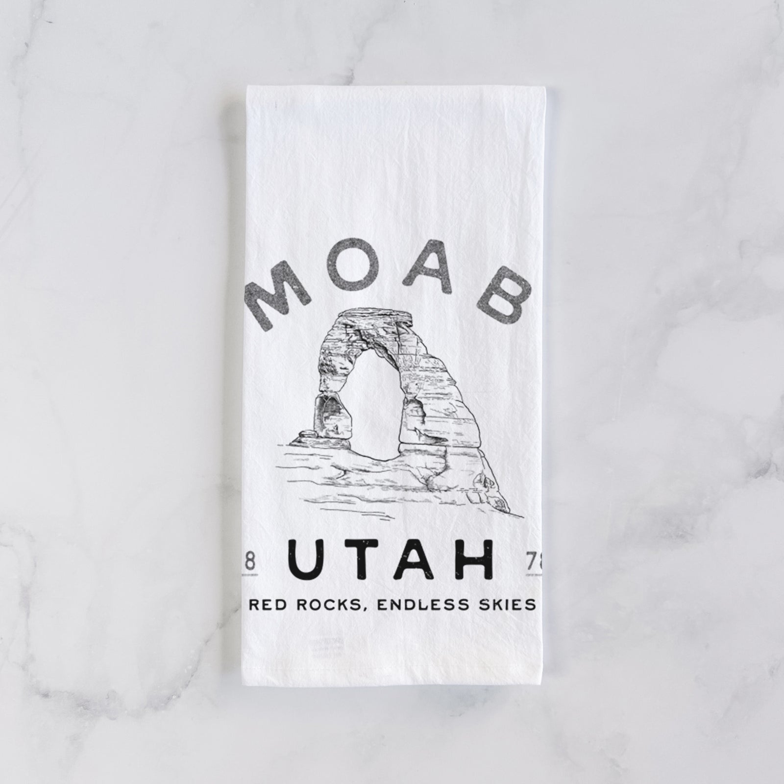 White cotton tea towel printed with a black ink illustration of Delicate Arch with the words Moab Utah, 1878, on a marble counter beside a round wooden cutting board and greenery.