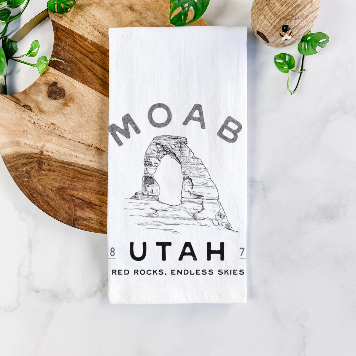 White cotton tea towel printed with a black ink illustration of Delicate Arch with the words Moab Utah, 1878, on a marble counter beside a round wooden cutting board and greenery.