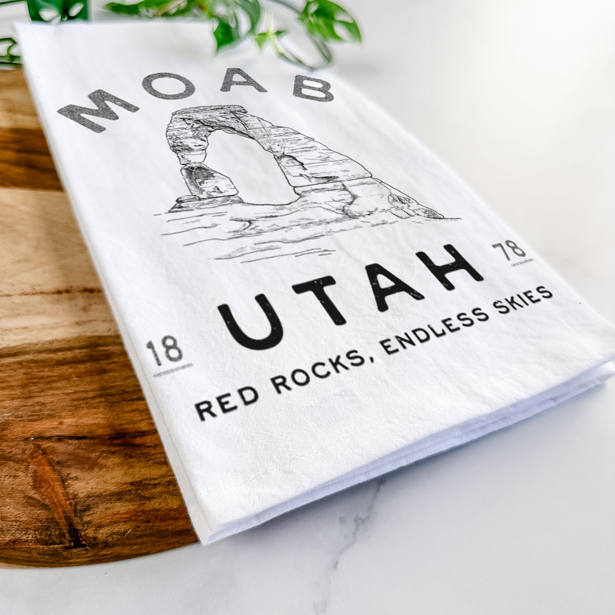 Close-up of a folded white tea towel on a wooden board, showing a black ink illustration of Delicate Arch with the words Moab Utah, 1878.