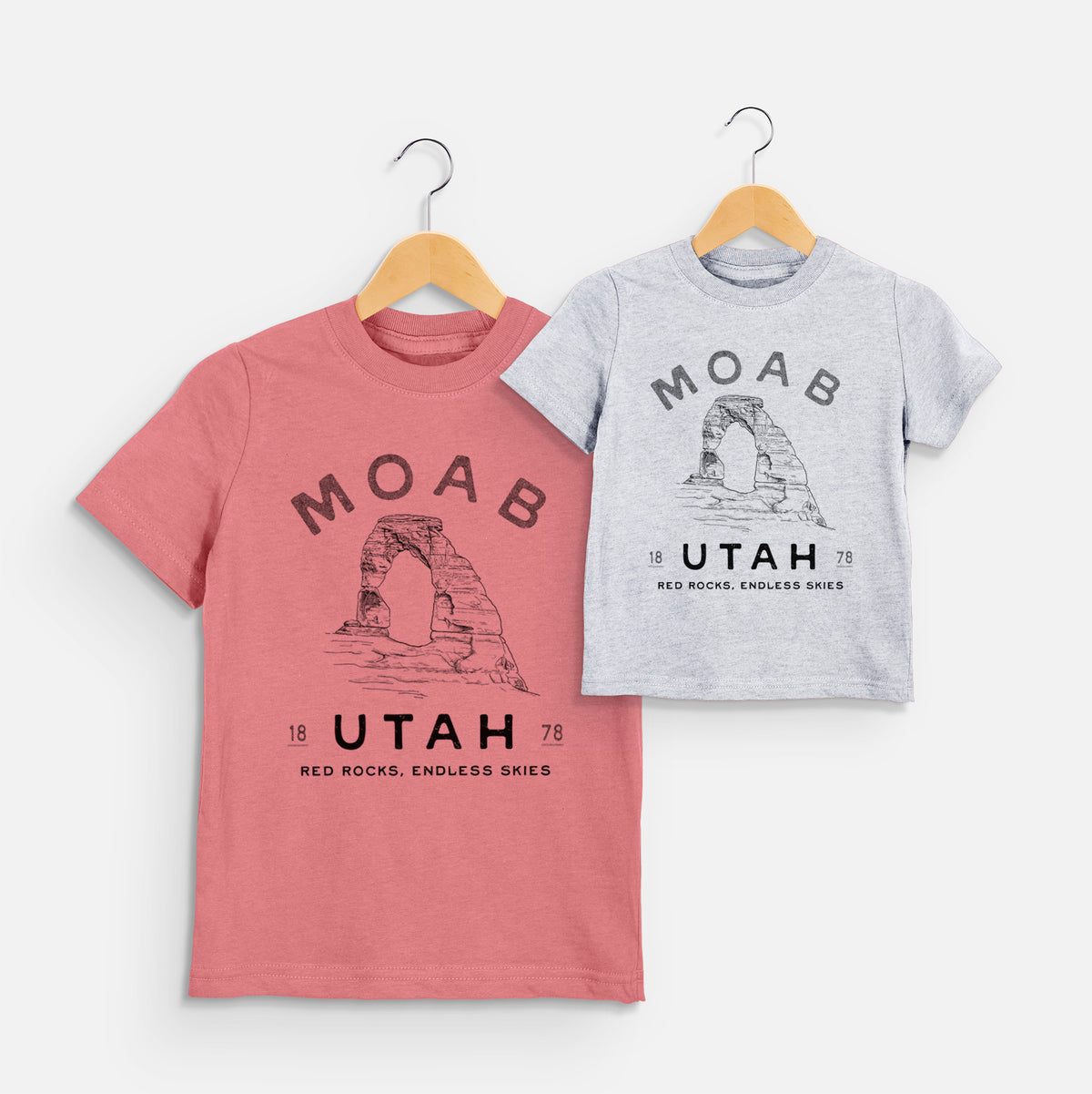 Two youth t-shirts in different colors on hangers, printed with a black ink illustration of Delicate Arch with the words Moab Utah, 1878.