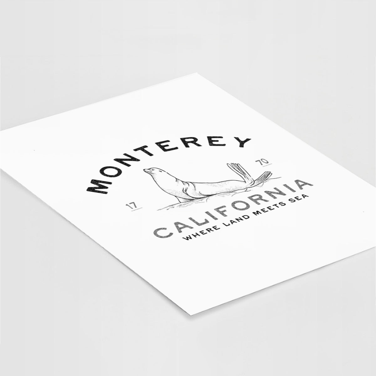 Art print of a black ink sea lion with the words Monterey California, 1770, Where Land Meets Sea, lying at an angle on a white surface.