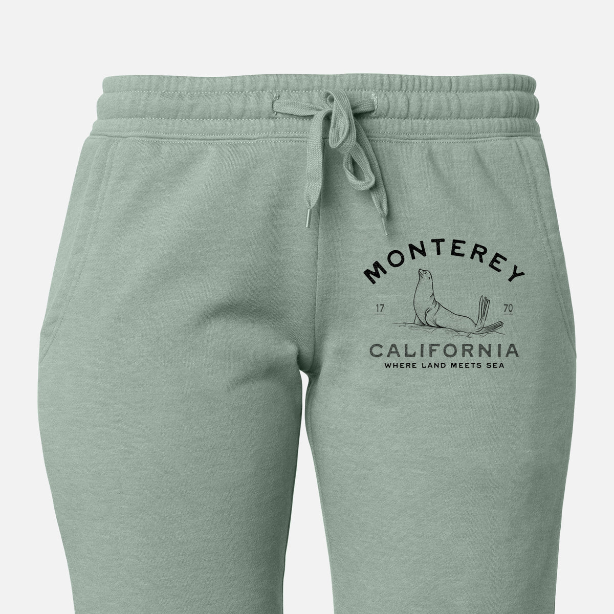 Front of sage green Cali Wave sweatpants, printed with a black ink sea lion with the words Monterey California, 1770, Where Land Meets Sea on the leg.