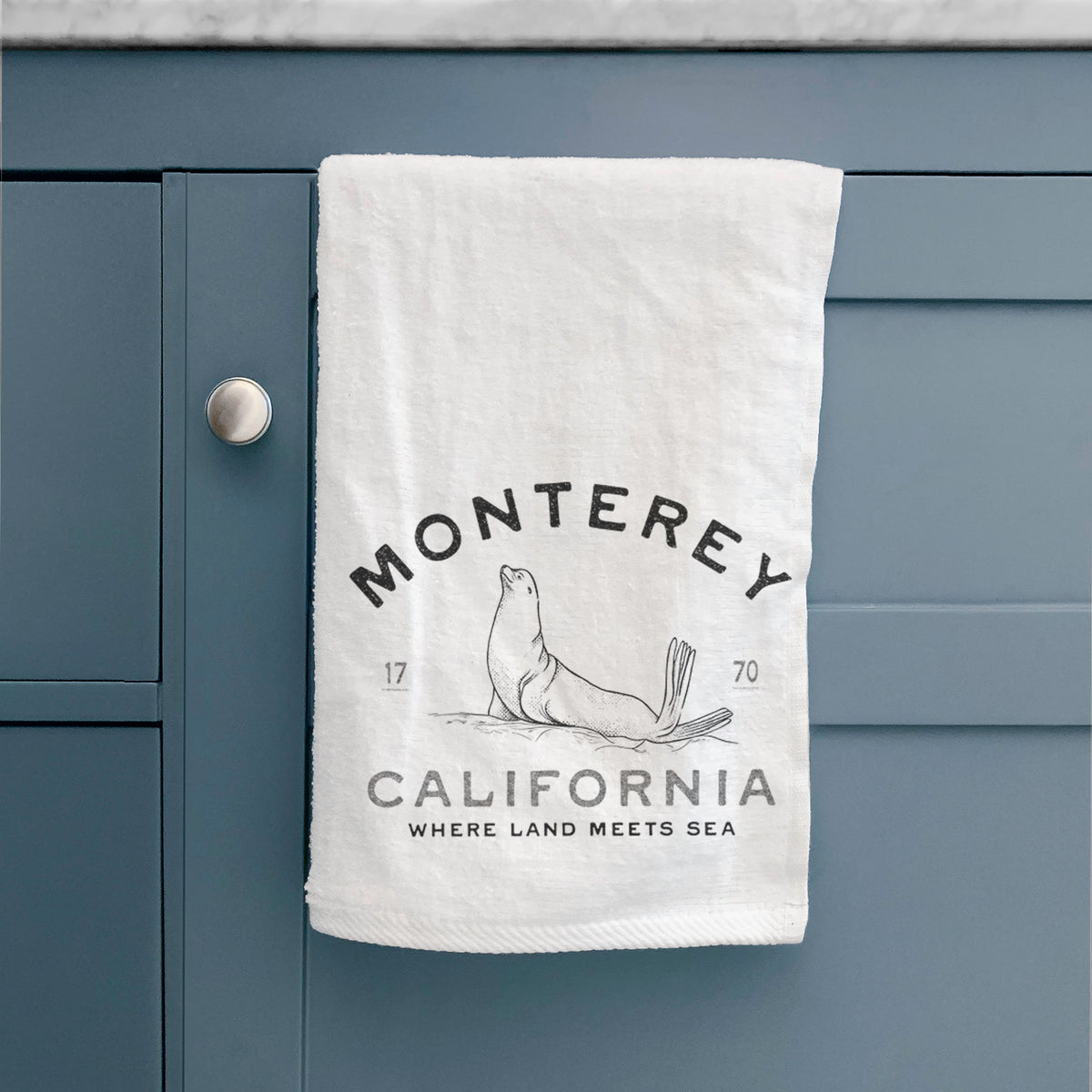 White hand towel printed with a black ink sea lion with the words Monterey California, 1770, Where Land Meets Sea, hanging on a cabinet door.