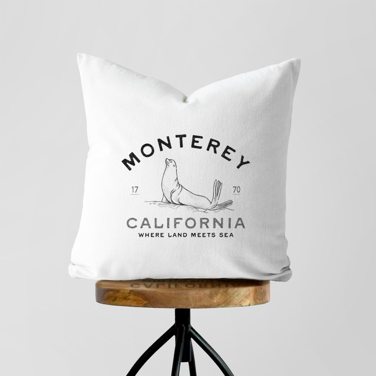 White pillow cover printed with a black ink sea lion with the words Monterey California, 1770, Where Land Meets Sea, on a wooden stool.